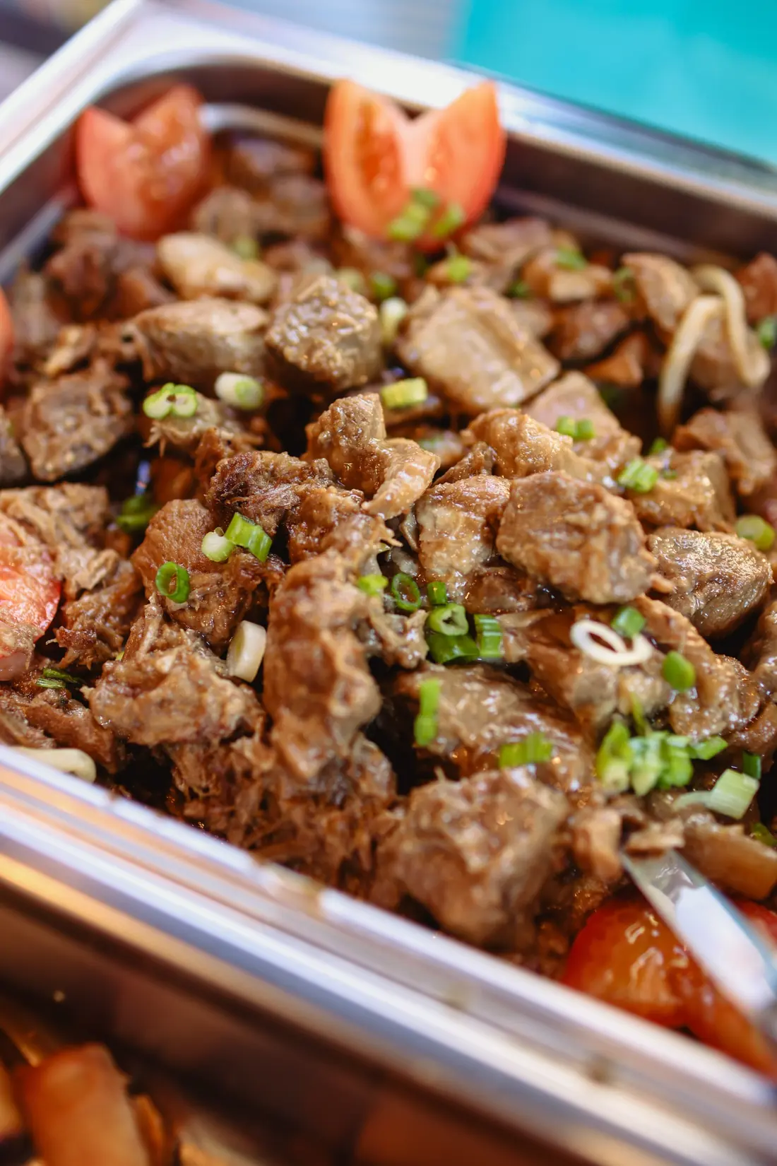 A chafing tray of braised meat garnished with tomato wedges and sliced green onions