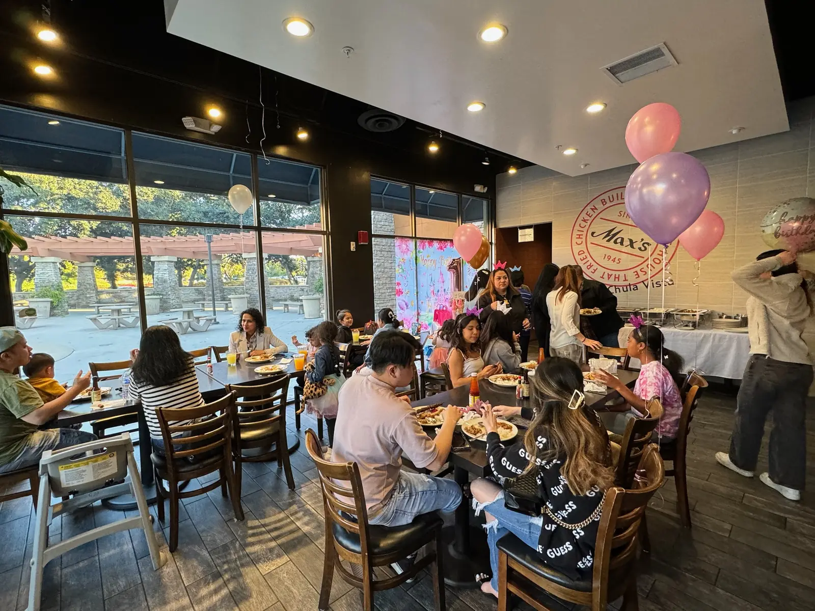 Family and friends in party crowns eat at tables by the windows during a first birthday at Max's Chula Vista, with pink and lavender balloons and the buffet by the Max's wall sign