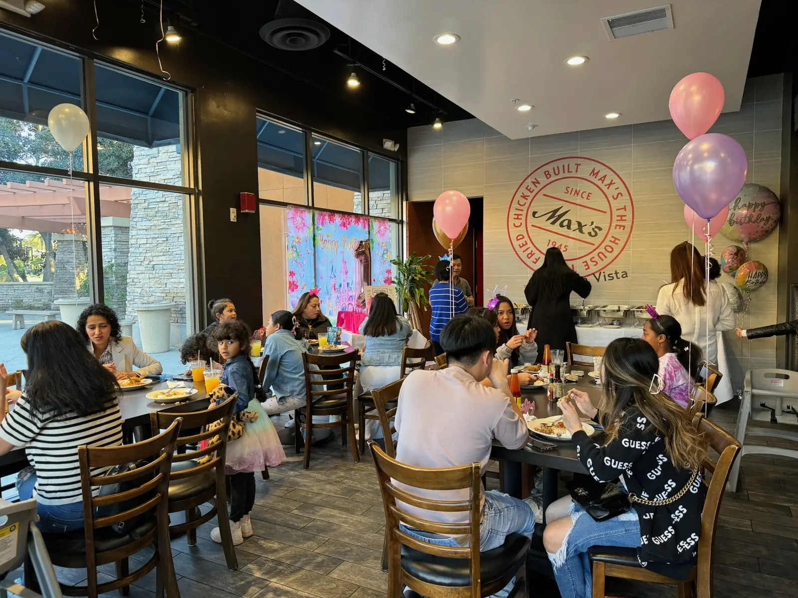 Guests of all ages eat at long tables under pink and lavender balloons at a first birthday at Max's Chula Vista, with the Max's Since 1945 wall sign behind them