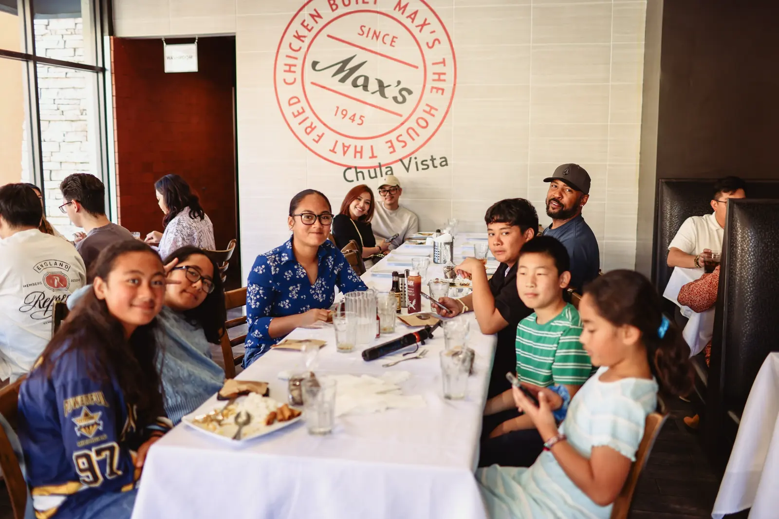 Family and friends of all ages around a long table in front of the Max's Since 1945 wall sign at Max's Chula Vista