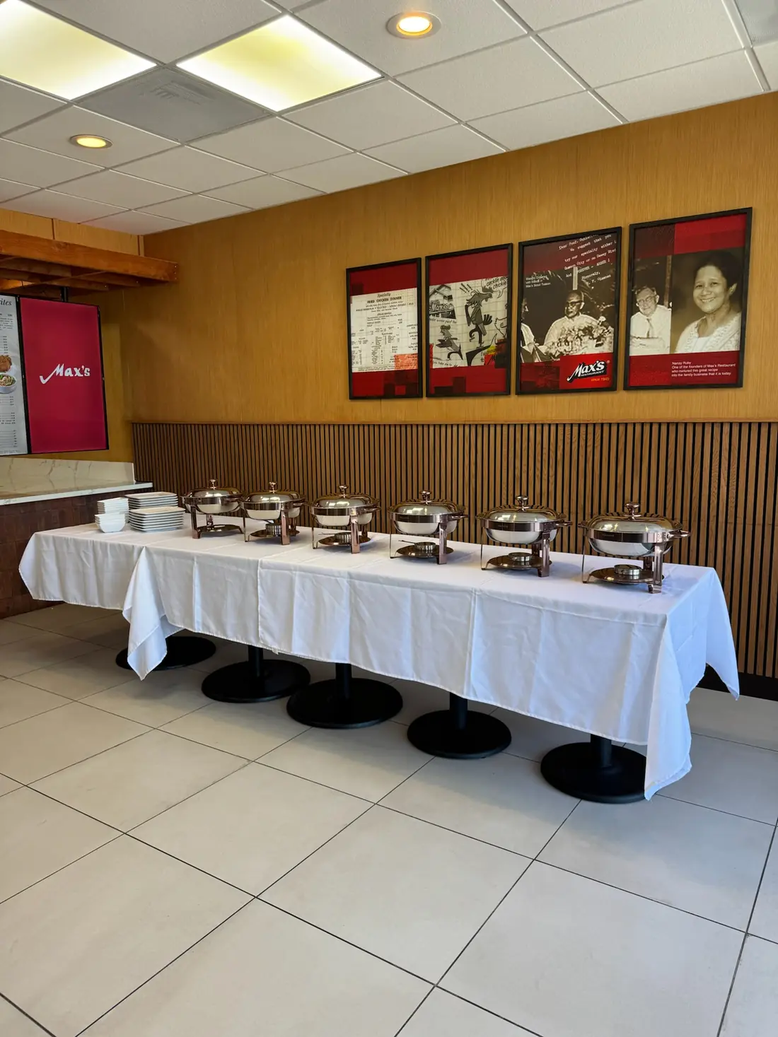 A buffet table with six copper-tone chafing dishes and stacked plates below framed photos of Max's founders at Max's National City