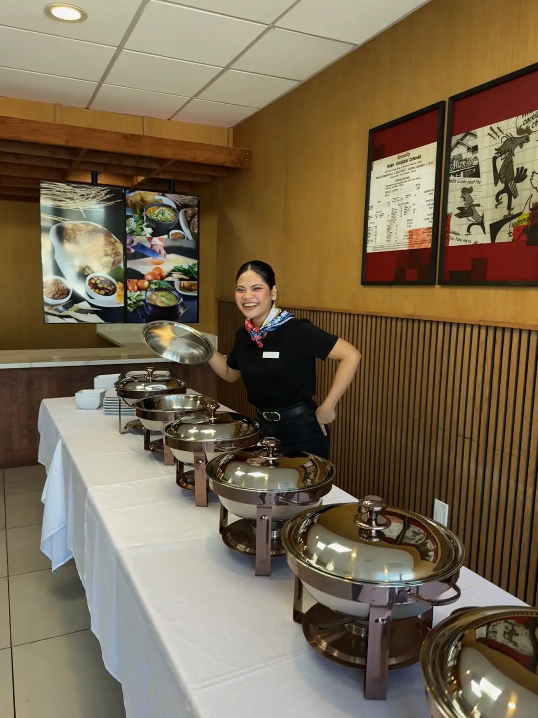 A smiling Max's host lifts the lid of a copper-tone chafing dish on the buffet table at Max's National City, below framed photos from Max's history