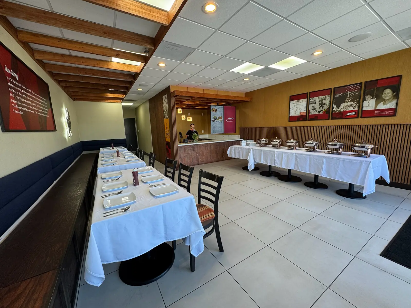 Long tables with white linens along a navy banquette, a buffet of copper-tone chafing dishes along the wood-slat wall and a host at the counter at Max's National City