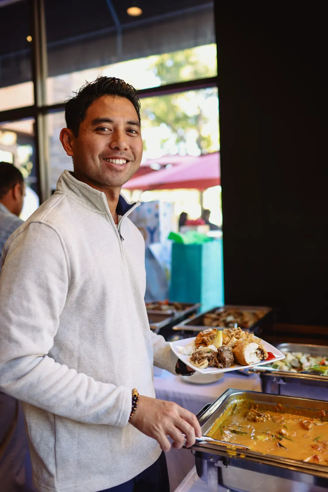 A smiling guest holds a full plate of Filipino party food next to a tray of kare-kare at a Max's Chula Vista buffet