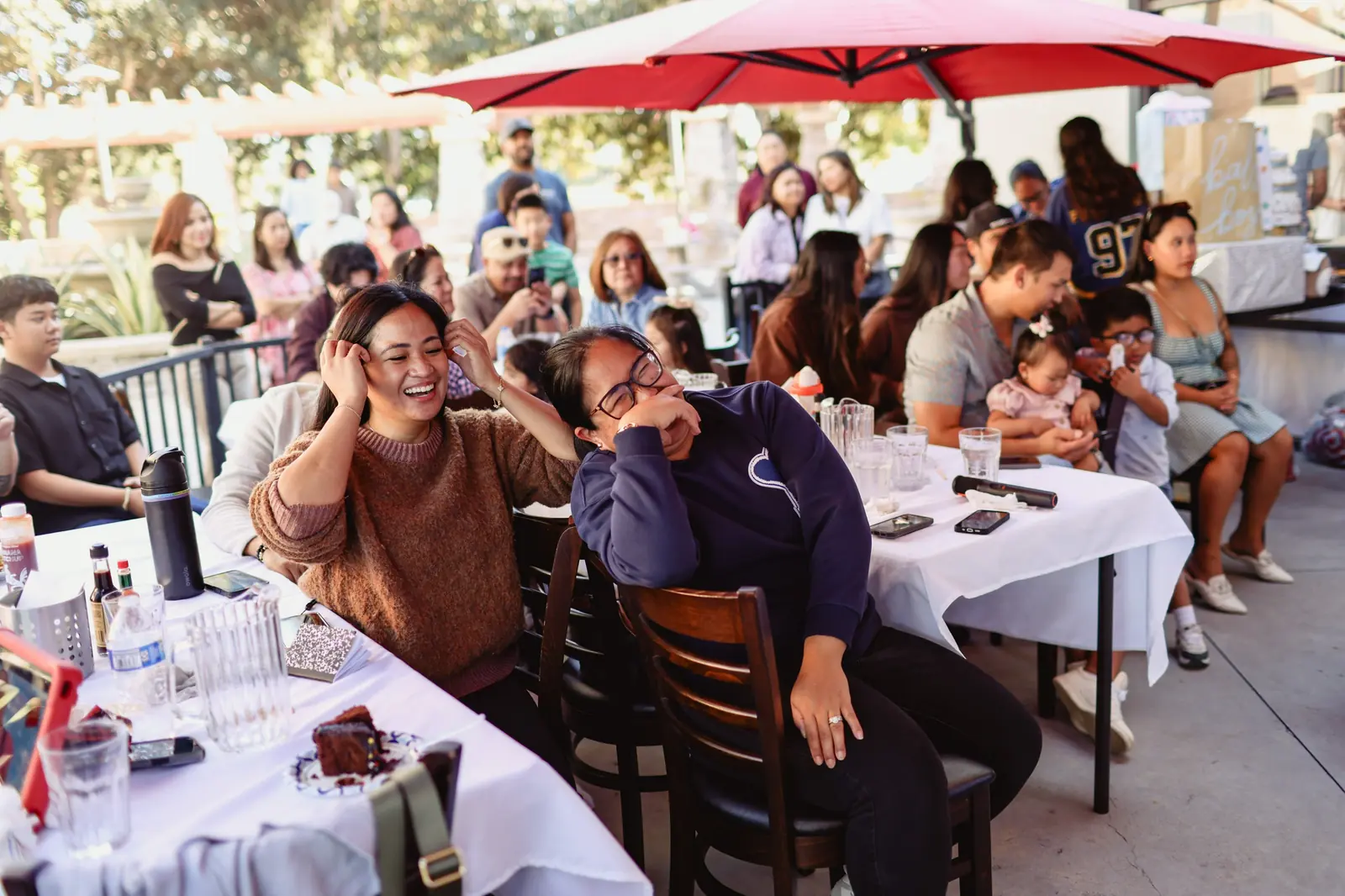 Two guests laugh together at a baby shower on the patio at Max's Chula Vista, with family seated under a red umbrella behind them