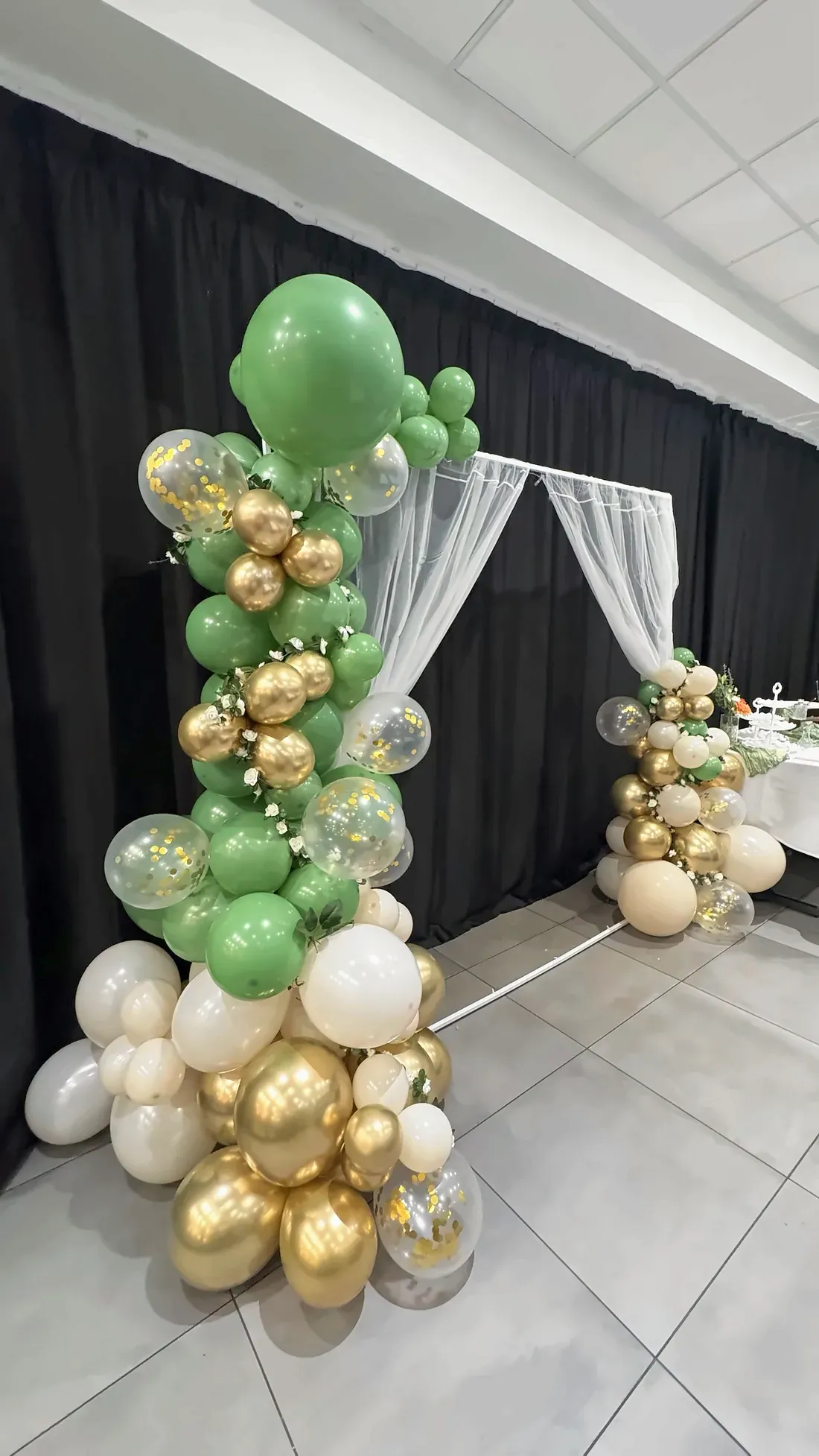 A green, gold and white balloon arch with a sheer white curtain, set against black drapes at Max's San Diego