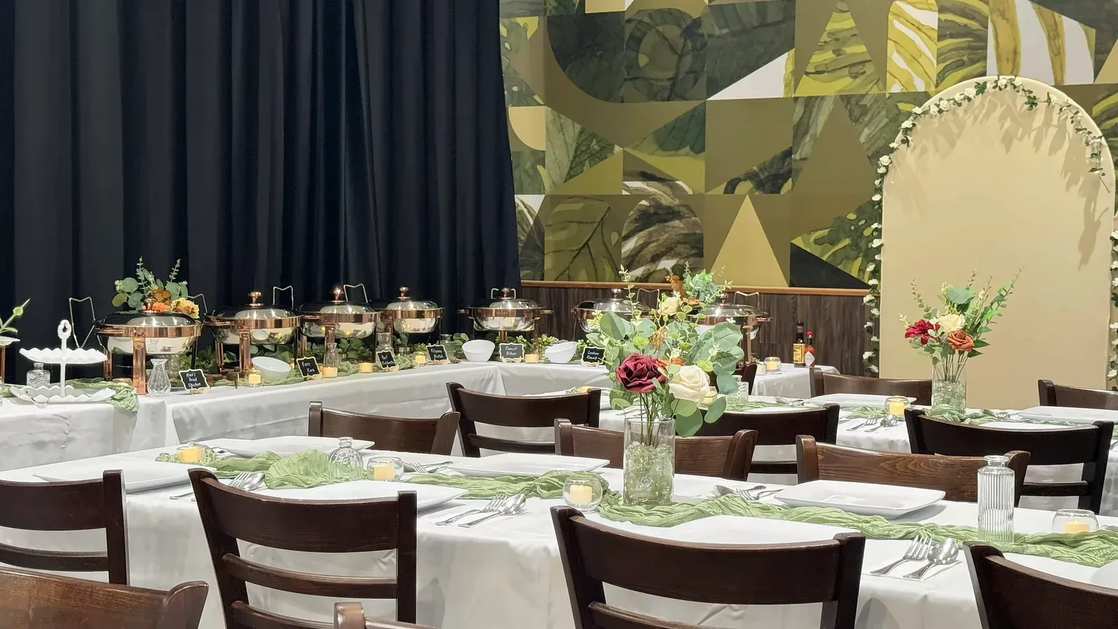 Long tables with white linens, green runners and flower centerpieces in front of a candlelit buffet of copper-tone chafing dishes and the leaf mural at Max's San Diego