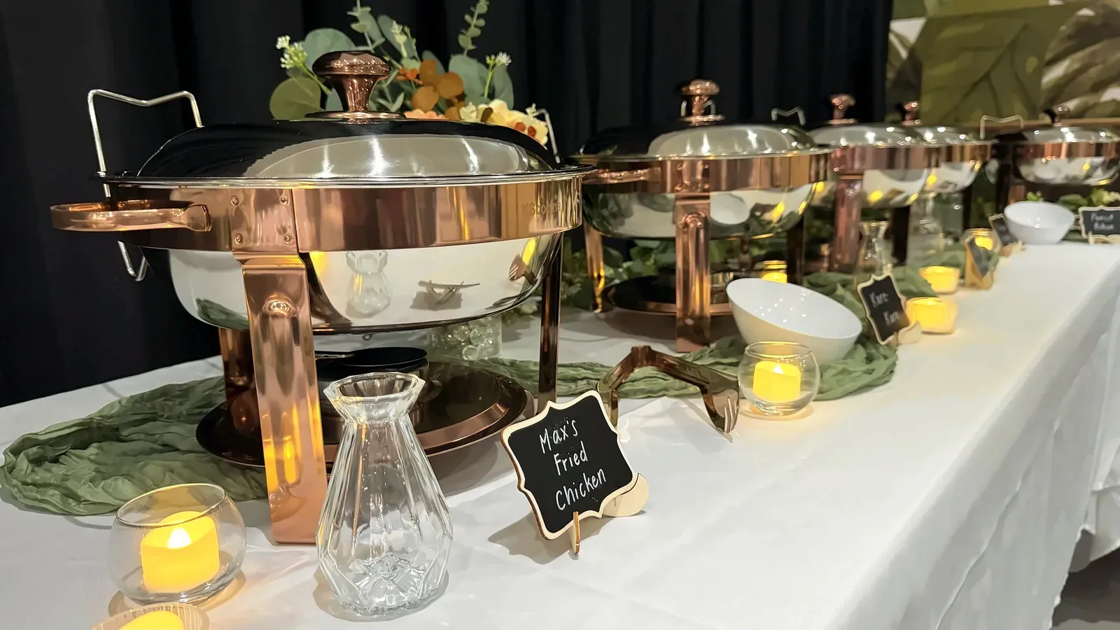 A line of copper-tone chafing dishes with chalkboard labels and candles along a white buffet table at Max's San Diego