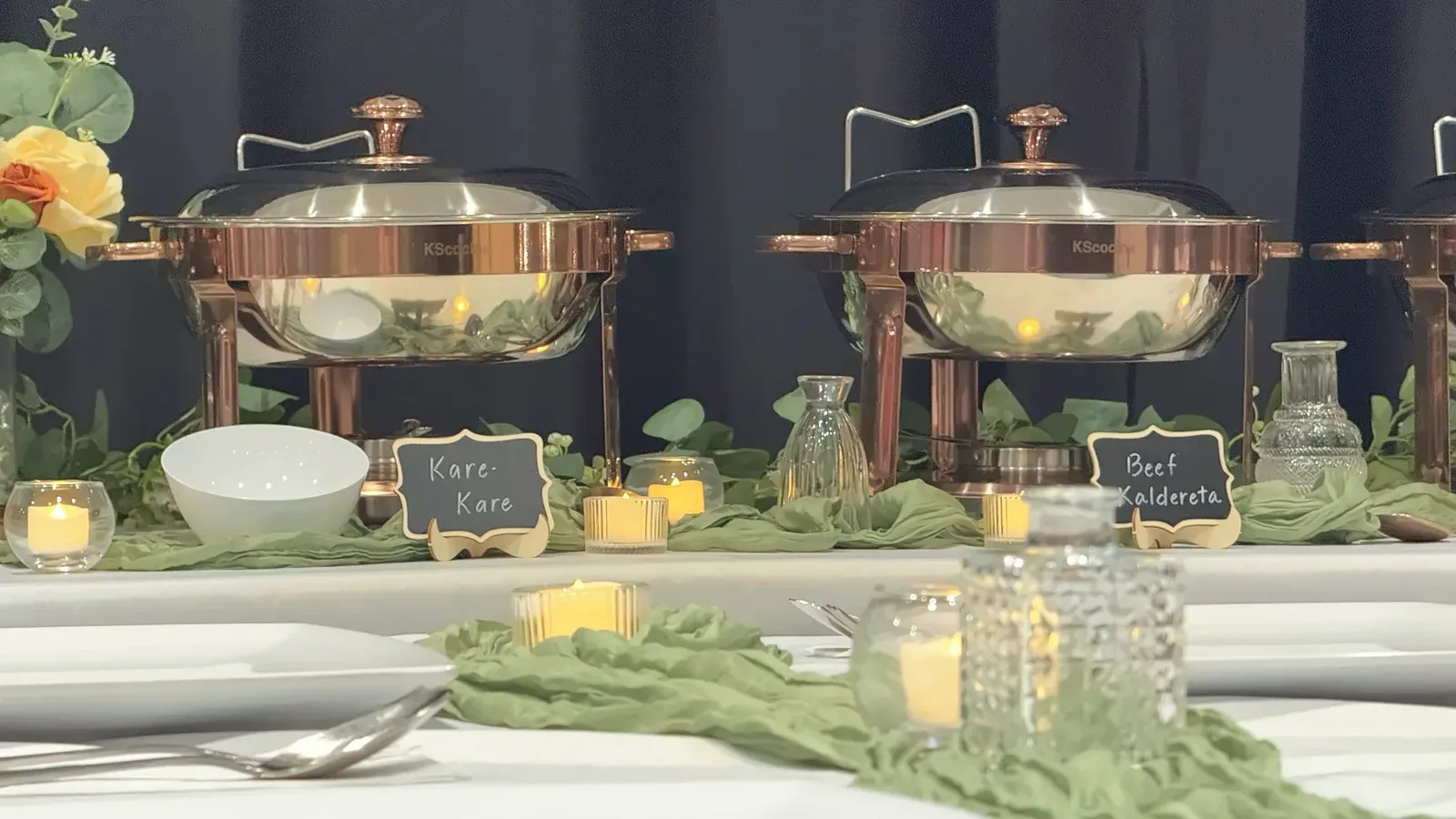Copper-tone chafing dishes labeled Kare-Kare and Beef Kaldereta with candles and greenery on the buffet at Max's San Diego