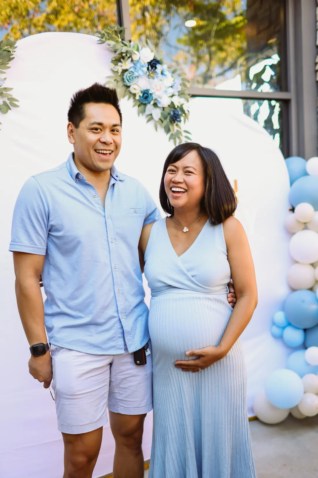 A smiling expecting couple in front of a floral and blue balloon backdrop at their baby shower at Max's Chula Vista